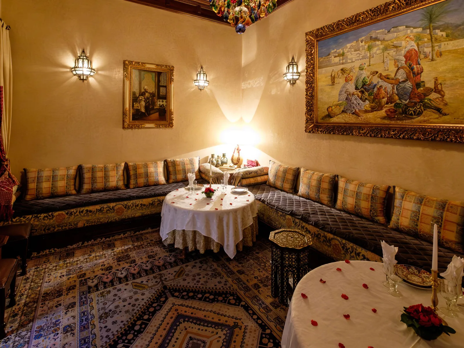 A Moroccan salon with built-in banquettes, a table set for dinner scattered with rose petals, rugs and a large painting